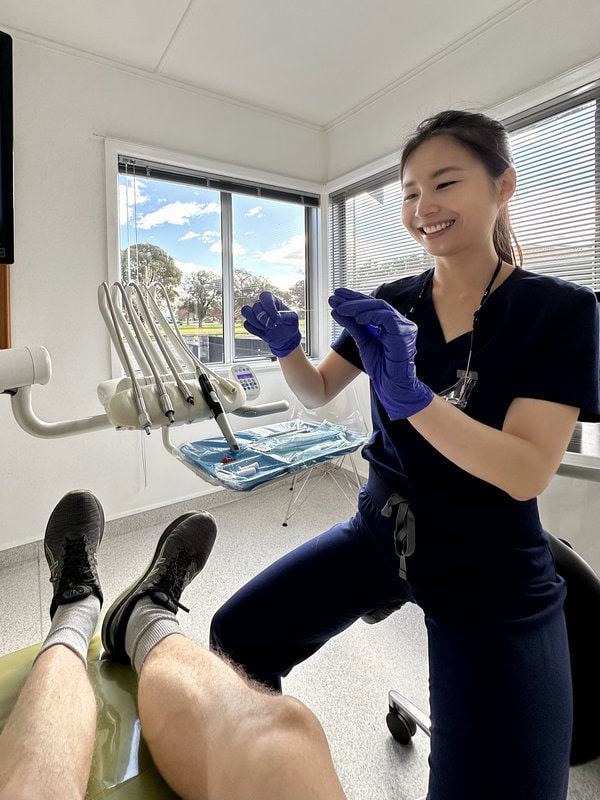 A dental professional wearing purple gloves prepares instruments while smiling at a seated patient in a bright dental office with large windows.